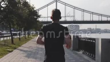 后侧跟随年轻活跃男子在城市码头沿河奔跑。 精力充沛的肌肉男孩跑得很快。 运动员跑步